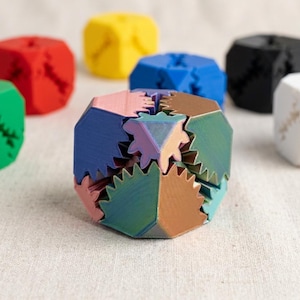 Könnte beinhalten: Eine Sammlung von mehrflächigen geometrischen Puzzlewürfeln mit ineinandergreifenden zahnradartigen Kanten. Der zentrale Würfel zeigt einen Farbverlauf aus Blau, Pink und Bronze, während die umliegenden Würfel einfarbig rot, gelb, blau, schwarz, grün und weiß sind.