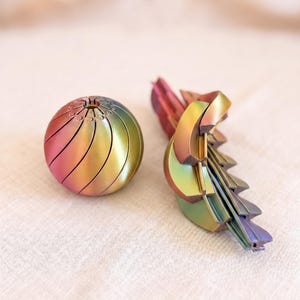 Könnte beinhalten: Ein kugelförmiges Objekt mit einer spiralförmig gemusterten Oberfläche und ein separates, längliches, bewegliches Teil, beide mit einer metallisch schillernden Oberfläche, die zwischen Rosa, Gold, Grün und Violett changiert.