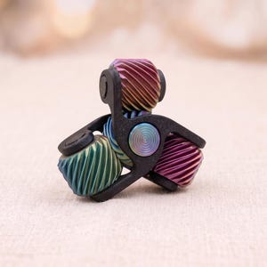 Könnte beinhalten: Ein Fidget Spinner mit einem schwarzen, dreizackigen Rahmen und einer zentralen, irisierenden runden Kappe. Jeder der drei Arme hält eine zylindrische Rolle mit spiralförmiger Rillenstruktur in den Farbverläufen Violett, Grün und Gold.