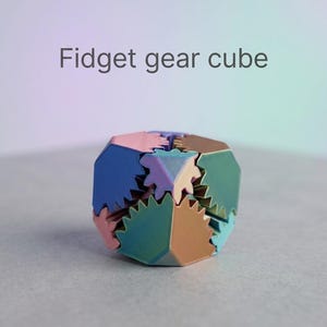 Könnte beinhalten: Ein mehrfarbiger Fidget-Zahnradwürfel mit ineinandergreifenden dreieckigen und zahnradförmigen Komponenten in Blau-, Rosa-, Grün- und Bronzetönen. Der Text "Fidget gear cube" ist in Grau über dem Objekt abgedruckt.