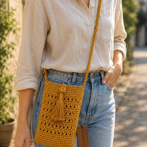 Peut inclure: Un sac bandoulière en crochet jaune moutarde avec un motif triangulaire ajouré et un pompon assorti. Le sac est doté d'une bandoulière tressée et d'une étiquette rectangulaire en similicuir marron attachée sur le côté avec du texte.