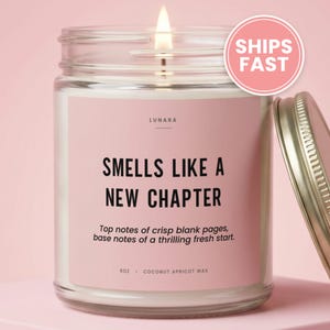 May include: A clear glass candle jar with a pink label featuring the text LUNARA, SMELLS LIKE A NEW CHAPTER, Top notes of crisp blank pages, base notes of a thrilling fresh start, 9oz, and COCONUT APRICOT WAX. A lit wick and a gold metal lid.