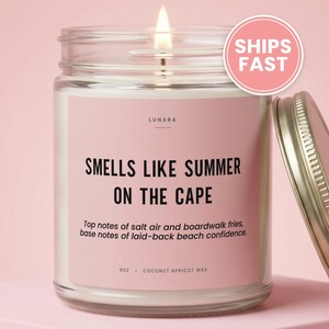 May include: A clear glass jar candle with a lit wick and a pink label. The label reads LUNARA, SMELLS LIKE SUMMER ON THE CAPE, Top notes of salt air and boardwalk fries, base notes of laid-back beach confidence, 9OZ, COCONUT APRICOT WAX. A metal lid sits nearby.