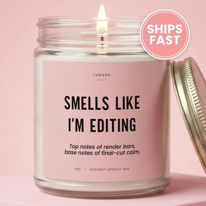 May include: A clear glass candle jar with a lit wick and a pink label. The label reads LUNARA, SMELLS LIKE I'M EDITING, Top notes of render bars, base notes of final-cut calm, 9oz, and COCONUT APRICOT WAX. A silver metal lid sits beside the jar.