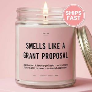 May include: A clear glass candle jar with a pink label featuring the text "SMELLS LIKE A GRANT PROPOSAL", "Top notes of freshly printed manuscripts, base notes of peer-reviewed optimism", "LUNARA", and "9oz - COCONUT APRICOT WAX". A lit wick is visible.