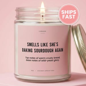 May include: A clear glass candle jar with a light pink label featuring the text "SMELLS LIKE SHE'S BAKING SOURDOUGH AGAIN" and "Top notes of warm crusty bread, base notes of wild-yeast glory." The candle contains 9oz of coconut apricot wax.