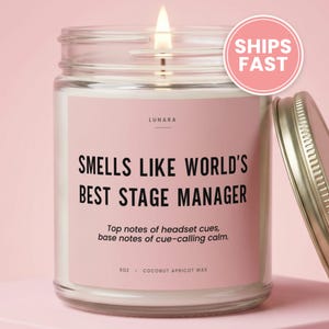 May include: A clear glass jar candle with a pink label featuring the text "SMELLS LIKE WORLD'S BEST STAGE MANAGER" and "Top notes of headset cues, base notes of cue-calling calm." The candle contains 9oz of coconut apricot wax and has a lit wick.