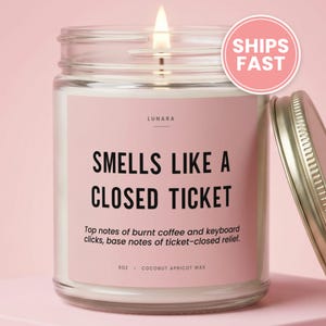 May include: A clear glass candle jar with a lit wick and a metallic lid. The pink label features the text LUNARA, SMELLS LIKE A CLOSED TICKET, and Top notes of burnt coffee and keyboard clicks, base notes of ticket-closed relief. It is 9oz.