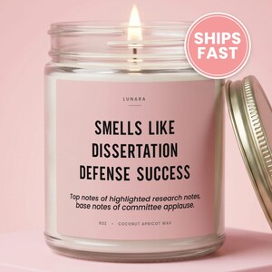 May include: A clear glass candle jar with a lit wick and a pink label. The label reads LUNARA, SMELLS LIKE DISSERTATION DEFENSE SUCCESS, Top notes of highlighted research notes, base notes of committee applause, 9oz, COCONUT APRICOT WAX. A metal lid sits nearby.