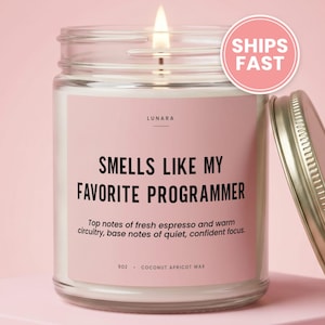 May include: A clear glass jar candle with a lit wick and a pink label. The label reads LUNARA, SMELLS LIKE MY FAVORITE PROGRAMMER, Top notes of fresh espresso and warm circuitry, base notes of quiet, confident focus, 9oz, COCONUT APRICOT WAX.