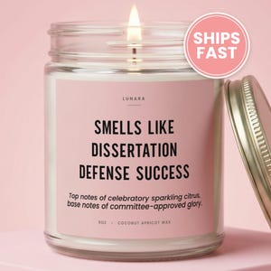 May include: A clear glass candle jar with a pink label featuring the text "LUNARA", "SMELLS LIKE DISSERTATION DEFENSE SUCCESS", and "Top notes of celebratory sparkling citrus, base notes of committee-approved glory. 9oz . COCONUT APRICOT WAX". The candle has a lit wick and a metallic lid.