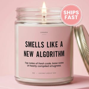 May include: A lit candle in a clear glass jar with a pink label. The label reads LUNARA, SMELLS LIKE A NEW ALGORITHM, Top notes of fresh code, base notes of freshly compiled smugness, 9oz, and COCONUT APRICOT WAX. A metal lid sits to the right.