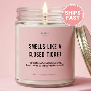 May include: A clear glass candle jar with a pink label featuring the text "LUNARA", "SMELLS LIKE A CLOSED TICKET", "Top notes of cooled circuitry, base notes of inbox-zero serenity", "9oz", and "COCONUT APRICOT WAX". A lit wick and a gold lid are visible.