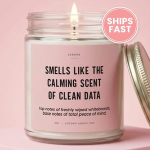 May include: A clear glass candle jar with a pink label featuring the text LUNARA, SMELLS LIKE THE CALMING SCENT OF CLEAN DATA, Top notes of freshly wiped whiteboards, base notes of total peace of mind, 9oz, and COCONUT APRICOT WAX. A metal lid sits beside.