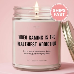May include: A clear glass candle jar with a lit wick and a pink label. The label reads LUNARA, VIDEO GAMING IS THE HEALTHIEST ADDICTION, Top notes of controllers, base notes of guilt-free playtime, 9oz, and COCONUT APRICOT WAX. A metal lid sits beside it.