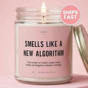 May include: A clear glass candle jar with a lit wick and a pink label. The label reads LUNARA, SMELLS LIKE A NEW ALGORITHM, Top notes of clean code, base notes of elegant solution energy, 9OZ, COCONUT APRICOT WAX. A metallic lid sits to the right.