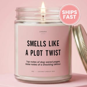 May include: A clear glass jar candle with a lit wick and a pink label. The label reads LUNARA, SMELLS LIKE A PLOT TWIST, Top notes of dog-eared pages, base notes of a shocking detour, 9oz, and COCONUT APRICOT WAX. A metallic lid sits to the right.