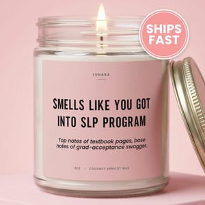 May include: A 9oz scented candle in a clear glass jar with a light pink label. The label reads: LUNARA, SMELLS LIKE YOU GOT INTO SLP PROGRAM, Top notes of textbook pages, base notes of grad-acceptance swagger, 9OZ, COCONUT APRICOT WAX. A lit wick.