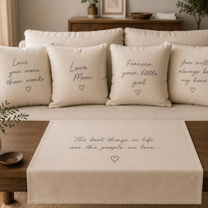 May include: Four beige square throw pillows and one rectangular table runner, all featuring black cursive text and a small heart icon. The pillows display phrases including Love you more than words, Love Mom, Forever your little girl, and You will always be my hero. The runner reads The best things in life are the people we love.