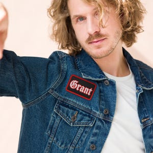 May include: A rectangular embroidered patch featuring the name Grant in white gothic-style lettering with a black background and a bold red border, attached to the chest pocket area of a dark blue denim jacket worn over a plain white t-shirt.