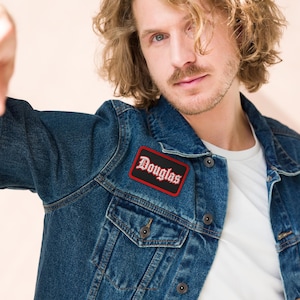 May include: A dark blue denim jacket featuring a rectangular embroidered patch on the chest. The patch has a black background with a red border and the name Douglas written in a white gothic-style font with a red outline.