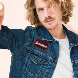 May include: A dark blue denim jacket featuring a rectangular patch on the chest. The patch has a black background with a red border and the name Robertson written in white gothic-style lettering across the centre of the fabric surface.