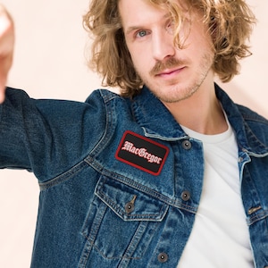 May include: A dark blue denim jacket featuring a rectangular patch on the chest. The patch has a black background with a red border and the text MacGregor written in a white gothic-style font across the centre of the fabric surface.