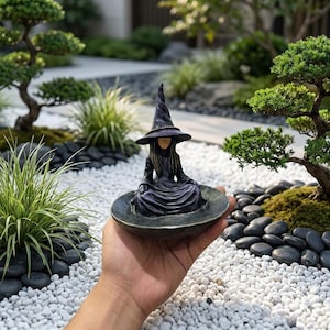 May include: A small figurine of a figure wearing a pointed witch hat and a long, flowing dark robe, seated in a meditative pose within a shallow, circular bowl-shaped base. The figure has long hair and a smooth, featureless face.