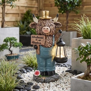 May include: A brown Highland cow figurine wearing a gold top hat and blue overalls, holding a wooden sign that reads Welcome to my Garden in one hand and a black lantern in the other, standing on a gray stone base with a small pink flower.