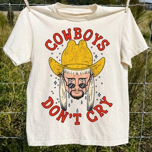 May include: A natural-colored short-sleeve t-shirt featuring a graphic print of a person wearing a yellow cowboy hat and sunglasses with long hair. The text "COWBOYS DON'T CRY" is printed in red above and below the illustration with blue teardrops.