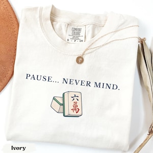 May include: An ivory short-sleeved t-shirt featuring the text PAUSE... NEVER MIND. in dark blue lettering above a graphic of two mahjong tiles. The shirt has a Comfort Colors brand label at the inner neckline and a thin gold chain necklace.