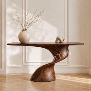 Puede incluir: Una mesa consola de madera marrón oscuro con una base escultural curva y lisa y una superficie plana. Sobre la mesa hay un jarrón de cerámica de color claro con ramas finas y desnudas y un pequeño objeto decorativo anudado.