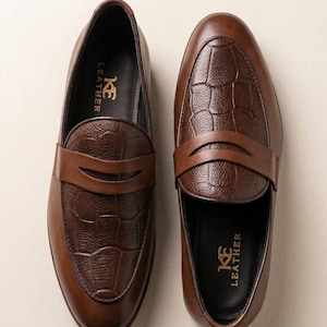 Può includere: Un paio di mocassini in pelle marrone con un motivo testurizzato a rilievo di coccodrillo sulla tomaia e una fascia in pelle liscia. La soletta interna nera presenta la scritta dorata KE LEATHER stampata nella zona del tallone.