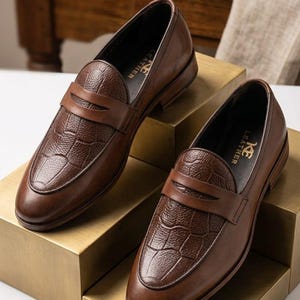 Könnte beinhalten: Ein Paar braune Leder-Loafer mit einer strukturierten Krokodilprägung auf dem Vorderblatt und einem klassischen Penny-Loafer-Steg. Die Innensohle ist mit dem Schriftzug KE LEATHER in goldener Schrift auf schwarzem Grund versehen.