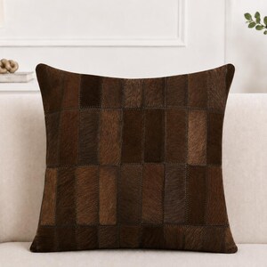 Puede incluir: Un cojín cuadrado con un diseño de patchwork compuesto por tiras rectangulares de piel animal en tonos marrón oscuro y marrón chocolate. La superficie muestra una textura natural de pelo sobre piel con costuras visibles uniendo cada panel.