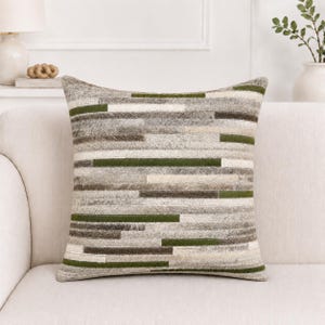 Puede incluir: Un cojín cuadrado con un diseño de retazos de tiras horizontales en tonos verde bosque, crema y varios matices de gris y marrón. La superficie tiene una textura similar a la piel con pelo, con costuras visibles entre cada tira.