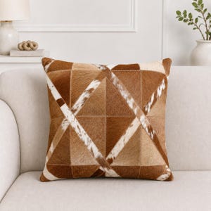 Puede incluir: Un cojín cuadrado con un diseño geométrico de patchwork hecho de piel de vaca. La superficie muestra un patrón de triángulos en varios tonos de marrón, bronceado y crema, con texturas de pelo sobre piel en blanco y marrón por toda la pieza.