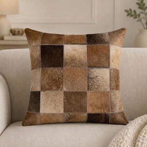 May include: A square throw pillow featuring a patchwork design of cowhide squares in various shades of brown, tan, and dark chocolate. The individual squares are stitched together with visible seams, creating a textured, multi-tonal surface.