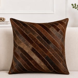Peut inclure: Un coussin carré présentant un motif en patchwork composé de bandes diagonales dans diverses nuances de brun. La surface a un aspect texturé de type peau avec poils, avec des coutures visibles entre chaque bande de matériau.