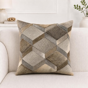 Könnte beinhalten: Ein quadratisches Zierkissen mit einem geometrischen Muster aus ineinandergreifenden Würfeln. Die Oberfläche besteht aus Fellstücken in verschiedenen Braun-, Beige-, Grau- und Cremetönen, was eine strukturierte, mehrdimensionale Optik erzeugt.
