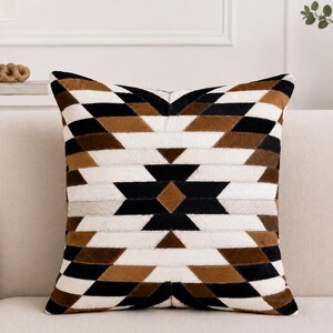 May include: A square throw cushion featuring a geometric pattern composed of stitched panels in black, white, and brown. The design consists of repeating triangles and diamond shapes arranged in a symmetrical, radiating layout across the surface.