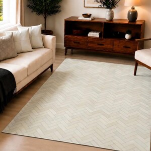 Puede incluir: Una alfombra rectangular con un patrón de espiga en tonos crema claro y blanco roto. La alfombra está colocada sobre un suelo de madera clara, cubriendo gran parte del espacio entre un sofá y un sillón en una sala de estar.