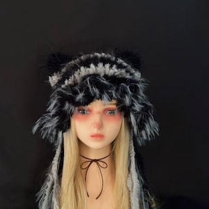 May include: A fuzzy, black and grey striped hat with ear-like protrusions on top and long, dangling side panels. The hat features a shaggy, textured faux fur material with a distinct pattern of alternating dark and light horizontal stripes.