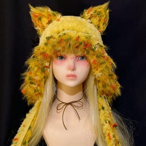 May include: A fuzzy yellow hat with pointed animal-like ears and long side flaps. The hat features a textured yarn blend with flecks of orange, green, and white throughout. A thin brown cord is tied in a bow around the neck of the mannequin.