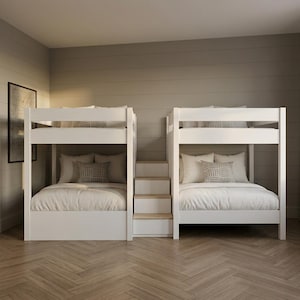 May include: Two white bunk beds connected by a central staircase with four steps. Each bed features a white frame, white bedding, and decorative pillows with a geometric pattern. The structure is positioned against a wall with horizontal paneling.
