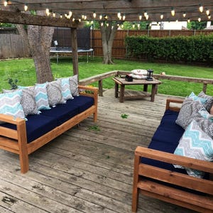 May include: Two wooden outdoor sofas with dark navy blue cushions and decorative pillows featuring gray and light blue chevron and floral patterns. The furniture is arranged on a wooden deck under a pergola with string lights in a backyard.
