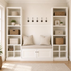 May include: A white entryway storage unit featuring two tall shelving towers flanking a central bench with two lower cabinets. The bench has a beige cushion and two throw pillows. Five dark metal hooks are mounted on the wall above the bench.