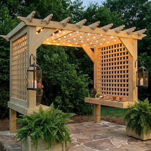 Op de afbeelding: Een houten pergola met een latwerk zijpaneel, een ingebouwde plank en twee zwarte metalen lantaarns met witte kaarsen. De balken zijn verlicht door een lichtsnoer, en er staan twee grote groene varens bij de constructie.