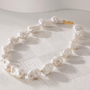Puede incluir: Un collar compuesto por numerosas perlas barrocas blancas, irregulares y brillantes, ensartadas en una disposición circular, con un pequeño cierre de metal dorado pulido que une los extremos del hilo de perlas de forma segura.