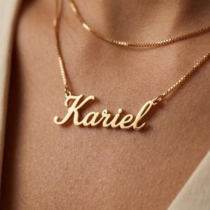 Puede incluir: Un collar de tono dorado con un colgante de placa con el nombre Kariel escrito en letra cursiva. El colgante está unido a una delicada cadena de eslabones cuadrados, con una segunda cadena dorada sencilla superpuesta encima.